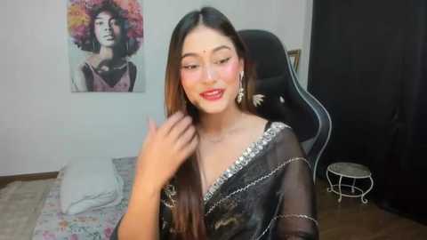 Media: Video of an East Asian woman with long dark hair, fair skin, wearing a black, sequined saree, red lipstick, and large earrings, sitting in a gaming chair in a modern bedroom.