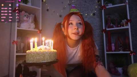 Video of a young woman with long, orange pigtails, wearing a green headband, and a white top, blowing out candles on a cake in a dimly lit room with shelves adorned with decorations.