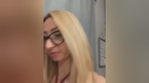 Media: A video of a blonde woman with long hair, wearing glasses and a red shirt, smiling slightly while looking at a person off-camera, in a blurred indoor setting.