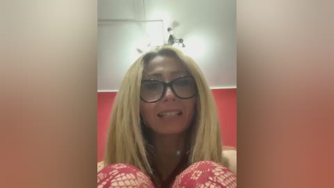 Media: Video of a smiling, blonde woman with large black glasses and a red patterned bra, set against a red and white wall with a ceiling fan.