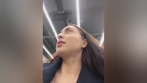 Video of a Caucasian woman with long brown hair, wearing a black top, gazing upwards in a modern, brightly lit room with recessed ceiling lights.