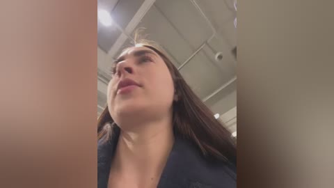 Video of a young woman with long brown hair, wearing a dark jacket, standing indoors with a ceiling fan visible, looking upward.