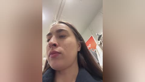 A close-up video of a young woman with long brown hair, fair skin, and freckles, making a pouty face. She wears a dark top in a store with beige walls, a red sign, and shelves in the background.