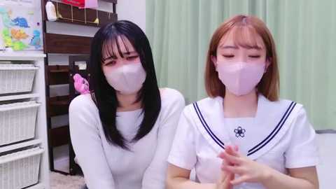 Media: Video of two young Asian women with light skin and straight black hair, wearing white school uniforms and masks, standing in a classroom with a map, shelves, and green curtains in the background.