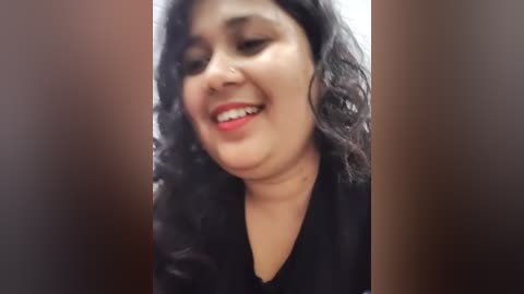 Media: Video of a smiling South Asian woman with curly black hair, wearing red lipstick and a black top, set against a blurred brown background.