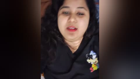 Media: Video of a young South Asian woman with curly black hair, light brown skin, wearing a black floral-patterned dress. She has a neutral expression. Background is dark.