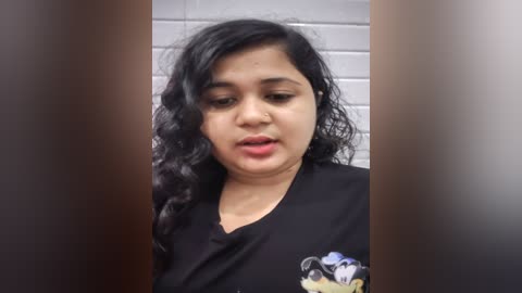 Media: Video of a young South Asian woman with curly black hair, wearing a black top, standing in a tiled bathroom. She has a solemn expression.