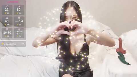 Media: A video of an East Asian woman with long black hair, fair skin, and slender build, wearing a black lace camisole, sitting on a white bed, making a heart shape with her hands, surrounded by sparkles.