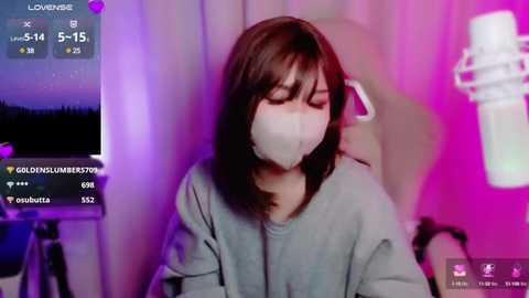 Media: Video of an Asian woman with shoulder-length brown hair, wearing a grey hoodie and white face mask, sitting in a room with pink and purple lighting. Live streaming on a computer screen.