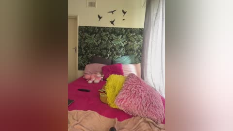 Video of a cluttered bedroom with a pink bedspread, fluffy yellow and pink pillows, a stuffed animal, and a jungle-themed wall.