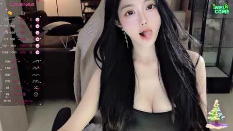 Media: A video of a young East Asian woman with long black hair, wearing a black tank top that reveals cleavage, sitting on a grey couch in a modern living room.