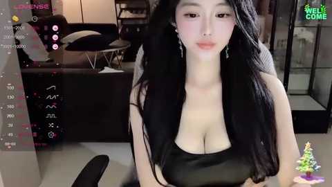 Media: A video of an East Asian woman with long black hair, fair skin, and large breasts in a black tank top, sitting in a modern living room with a TV screen in the background.