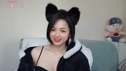 Media: Video of a young Asian woman with short black hair, wearing a black hoodie with white fur trim and cat ears, smiling, sitting on a sofa with a plush toy in the background.