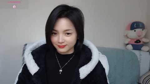 Media: A video of an Asian woman with straight black hair, fair skin, and red lipstick, wearing a black hoodie with white fur trim, seated on a blue couch. Background includes a plush toy and a pink water bottle.