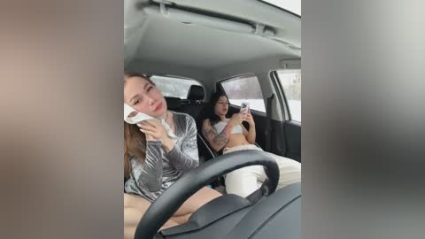 Video of two women in a car, one with a white bandage on her face, the other with a tattoo sleeve, wearing silver glittery tops and white pants.