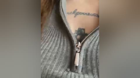 Media: A close-up video of a woman's chest in a gray zip-up sweater, revealing a tattoo of cursive text on her neck. The background is blurred and neutral-colored, focusing on the tattoo and clothing texture.