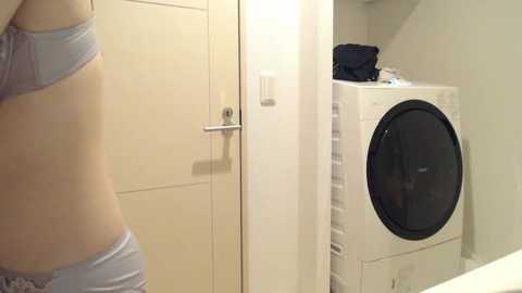 Media: Video of a woman in light blue underwear, side profile, standing near a white front-loading washer in a beige-tiled bathroom.