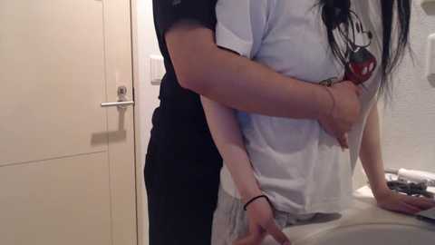 Media: A video of a woman in a white T-shirt, being embraced from behind by a man in a black shirt. The setting is a bathroom with beige tiles and a white sink.