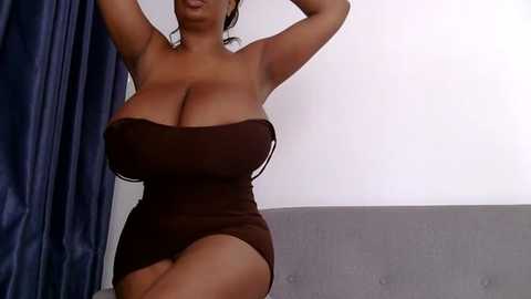 Media: Video of a voluptuous, dark-skinned woman with large breasts, wearing a tight, strapless brown dress, posing with arms raised, against a plain white wall and blue curtains.