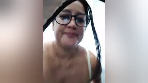 Media: Video of a smiling woman with glasses, fair skin, and long black hair, wearing a white bra, looking through a blurred window.