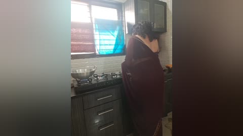 Media: Video of a woman in a maroon apron standing in a kitchen with dark wood cabinets, white tiled backsplash, and a blue window.