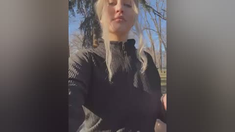 Media: A video of a blonde woman with fair skin wearing a black, ribbed, high-necked sweater, posing outdoors with a slight pout. Background features leafless trees and a clear blue sky.