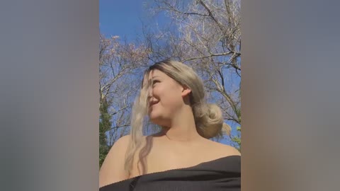 Media: Video of a smiling young woman with fair skin, blonde hair styled in a bun, wearing a black off-shoulder dress, standing outdoors with a clear blue sky and bare trees in the background.