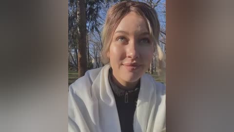 Media: A video of a fair-skinned, blonde woman with green eyes, wearing a white robe over a black turtleneck, standing in a sunlit forest with tall trees.