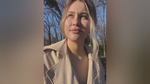 Media: Video of a smiling Caucasian woman with light blonde hair, wearing a beige trench coat, standing outdoors in a sunlit forest with bare trees and green foliage.