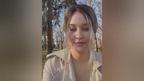 Media: Video of a young Caucasian woman with fair skin, light brown hair, and light makeup, wearing a beige hoodie, standing in a sunlit park with trees in the background.