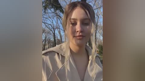 Media: Video of a fair-skinned woman with blonde hair, wearing a beige jacket, standing in a forest with leafless trees and a clear blue sky.