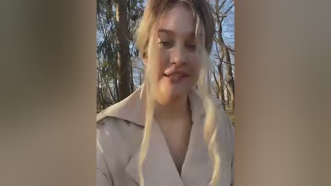 Media: Video of a smiling young woman with light skin and blonde hair, wearing a beige coat, standing in a forest with tall trees and a clear sky.