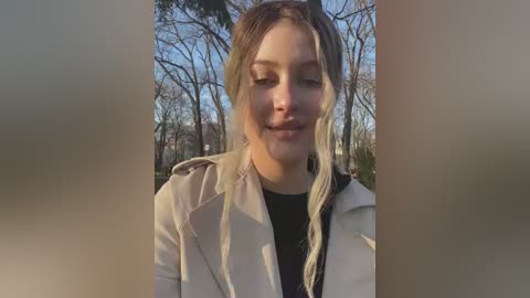 A video of a young woman with fair skin and light brown hair in a braid, wearing a beige coat and black top, smiling in a park with bare trees and a clear blue sky in the background.