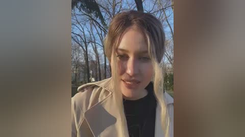 Media: Video of a smiling Caucasian woman with light brown hair, wearing a beige jacket and black shirt, outdoors in a forest with bare trees and blue sky background.