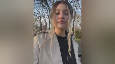 Media: A video of a young woman with long, blonde hair in a light beige jacket, standing outdoors in a park with bare trees in the background. She has a neutral expression.