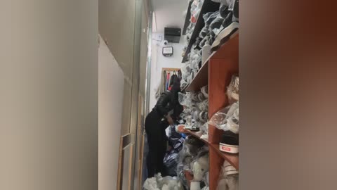 Media: Video of a narrow, cluttered hallway with a person in dark clothing and a black backpack, surrounded by stacks of white plastic bags, shelves filled with more bags, and a small office space visible in the background.