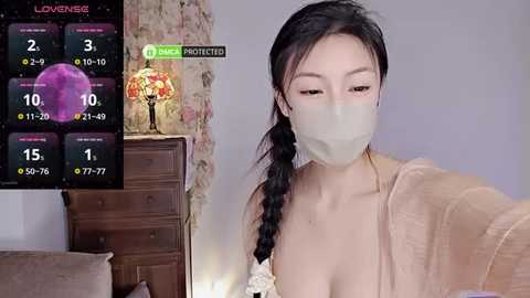 Media: Video of a young Asian woman with light skin, wearing a beige mask, braided black hair, and a beige robe, standing indoors.