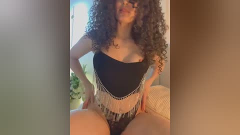 Media: Video of a woman with curly dark hair, wearing a black off-shoulder top with silver fringe and black panties, seated on a beige couch, indoors.