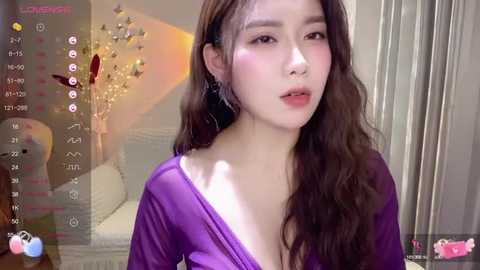 Media: A video of an Asian woman with long, wavy brown hair and fair skin, wearing a deep purple, off-shoulder dress, in a modern, softly lit room with a chandelier and decorative elements.