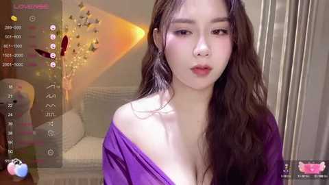 Media: Video of an East Asian woman with long brown hair, wearing a purple off-shoulder top, standing indoors. Background features a cozy, dimly lit room with a white armchair and decorative lights.