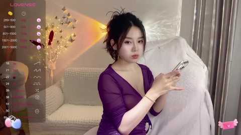 Media: A video of an East Asian woman with fair skin, dark hair tied back, wearing a purple robe, holding a needle and thread in a modern, light-colored room with a white couch and Christmas decorations.