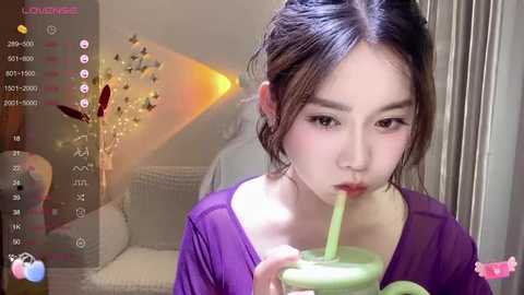 Media: A video of an East Asian woman with dark hair, wearing a purple top, drinking green juice from a straw, in a cozy room with a lit candle and a wicker chair.