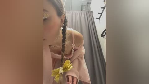 Media: Video of a young girl with long, braided hair adorned with yellow flowers, wearing a pink dress, looking at herself in a mirror, standing in a dimly lit dressing room with a grey curtain in the background.