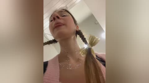 Media: Video of a young woman with long, braided hair, wearing a pink cardigan, captured from a low angle, showing her neck and shoulders. The background features a light-colored ceiling and a slightly blurred room.