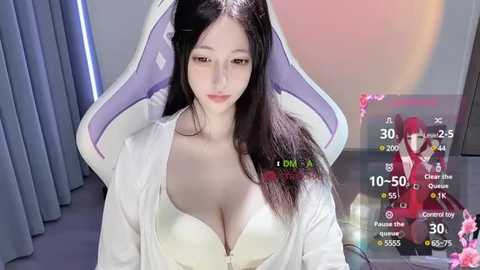 Media: A video of an East Asian woman with long black hair, fair skin, and large breasts, wearing a white robe, seated on a purple gaming chair. Background includes blue curtains and a virtual game interface.