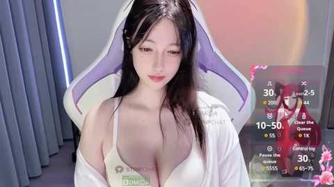 Media: Video of an East Asian woman with long black hair, wearing a white hoodie with \"Bikini\" written on it, in a modern room with blue curtains and a gaming monitor showing stats.