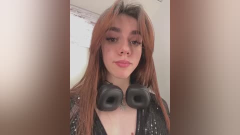 Media: A video of a young woman with long, straight reddish-brown hair, wearing a black sequined top and large black over-ear headphones. She has fair skin and a neutral expression. The background is blurred and neutral-toned.