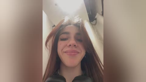 Media: Video of a young woman with long, dark hair, smiling broadly, with a light complexion. She's in a public bathroom, with white walls and a ceiling light visible.