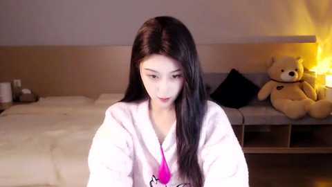 Media: A video of an East Asian woman with long black hair, fair skin, and petite frame, wearing a white robe, sitting in a modern bedroom with a beige bed, teddy bear, and warm lighting.