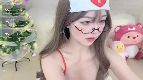 Media: A video of an East Asian woman with long brown hair, wearing a white nurse cap, red spaghetti straps, and black-rimmed glasses, sitting next to a decorated Christmas tree and plush toys in a cozy room.